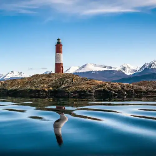 El faro en una isla con montañas nevadas al fondo y reflejo en el agua.