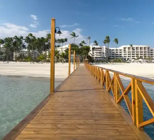 A wooden pier extending over clear blue water towards a beachfront with modern buildings and palm tr.
