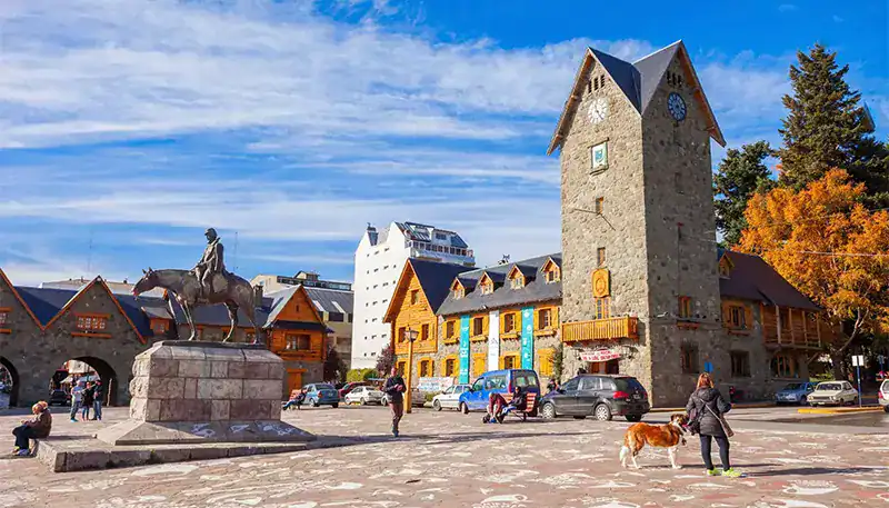 Historic stone building with clock tower in a lively town square, autumn trees, and people walking w.