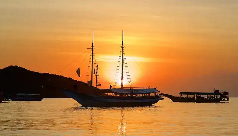 Barco de vela al atardecer en el puerto, vista panorámica con cielo naranja y reflejo en el agua.
