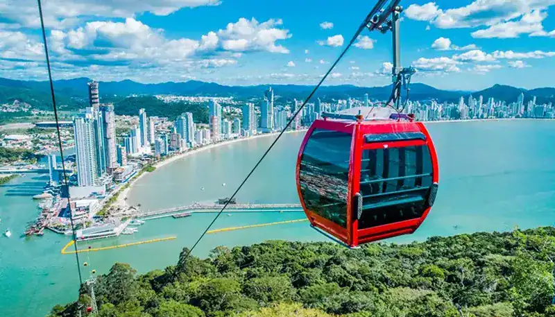 Aerial view of a red cable car over a coastal city skyline and lush green hills.