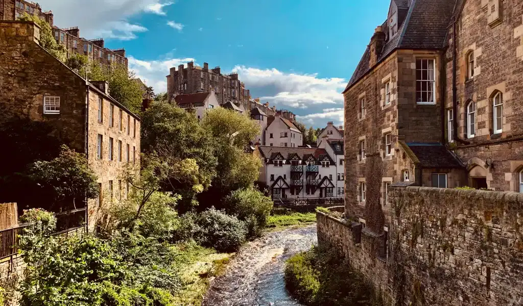 Puente antiguo sobre río en un pueblo europeo con casas de piedra y vegetación exuberante.