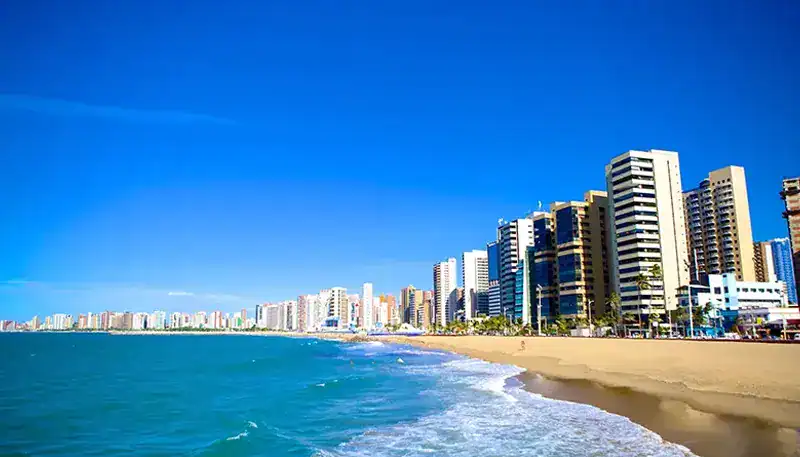 Bright city skyline along a sandy beach with clear blue sky and calm ocean waters.