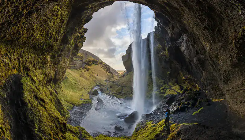 Cascada en una cueva natural con vegetación y paisaje montañoso al fondo.
