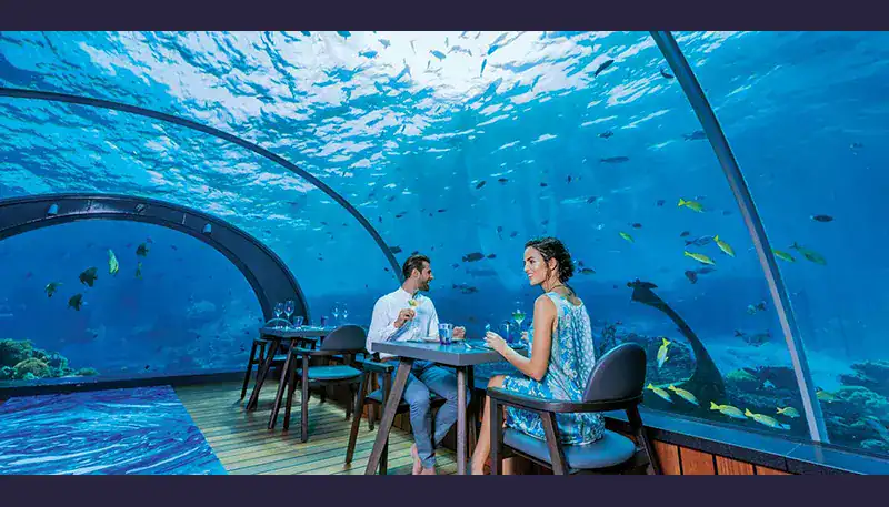 Elegant couple dining inside an underwater restaurant with large glass windows showcasing marine lif.