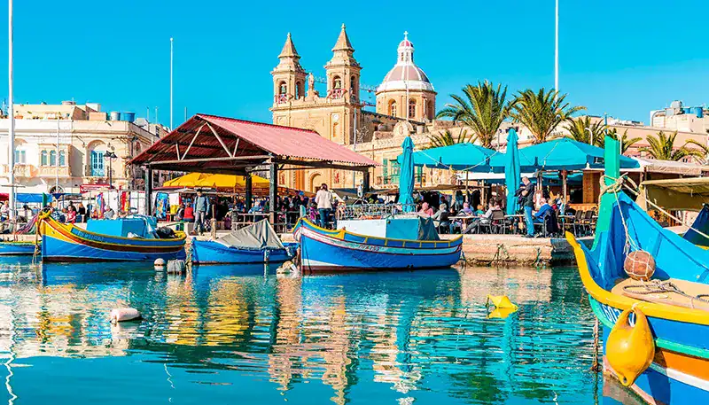 Colorful boats docked at a lively harbor with historic buildings and palm trees in the background.