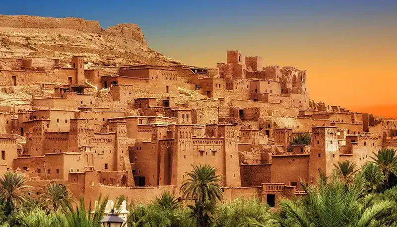 Ancient desert fortress with mud-brick buildings at sunset, surrounded by palm trees.