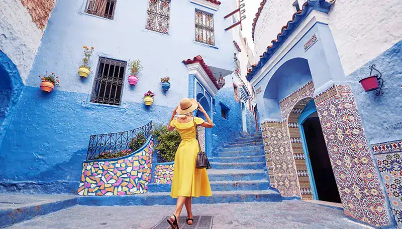 A woman in a yellow dress and hat walking through a colorful, vibrant street with blue walls and dec.