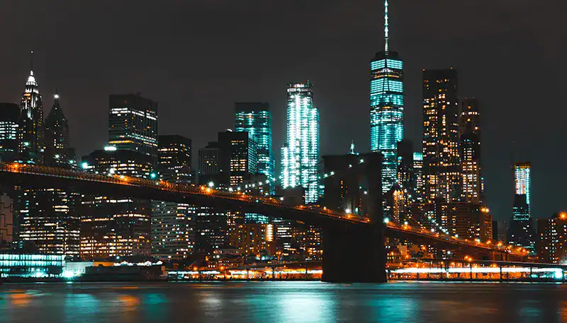 A nighttime view of New York City skyline with illuminated skyscrapers and Brooklyn Bridge.