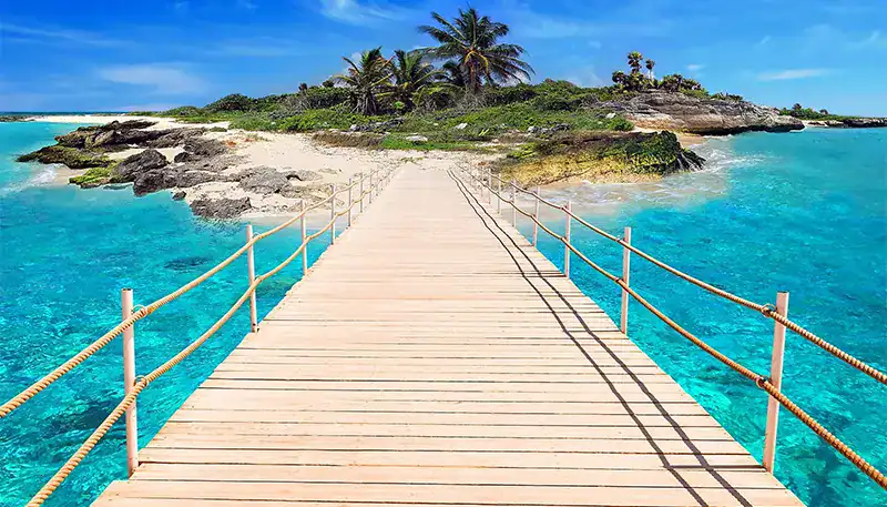 A wooden pier extending over turquoise waters towards a small island with palm trees.
