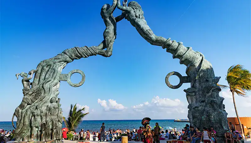 Colorful sculpture of two intertwined trees with rings, set against a beach background with people a.