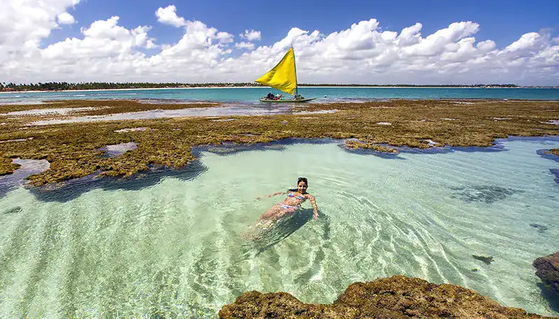 A beach scene with a woman swimming in clear shallow water and a sailboat with a yellow sail in the.