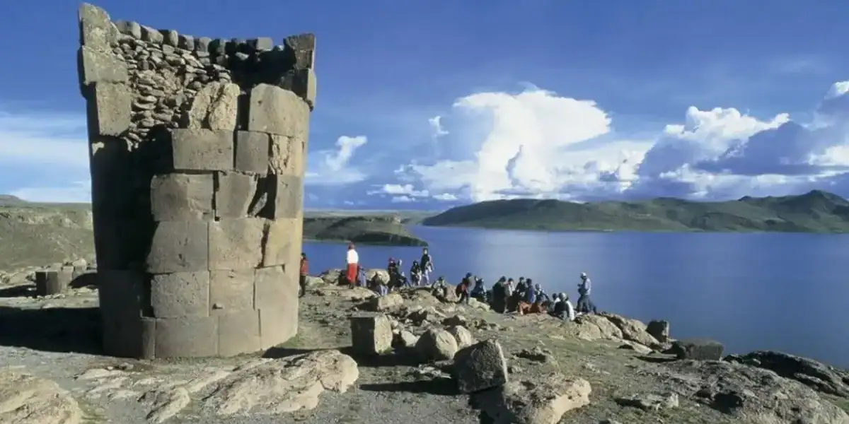Ancient stone structure by a lake with people enjoying the view and mountains in the background.