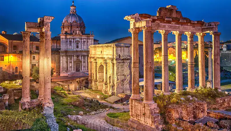 Ancient Roman ruins with illuminated historical buildings at dusk in Rome, Italy.