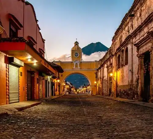 A street view of Antigua Guatemala with colonial architecture and a prominent clock tower at sunset.