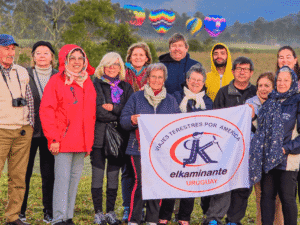Viajantes con bandera de El Kaminante en un campo abierto con globos aerostáticos en el cielo.