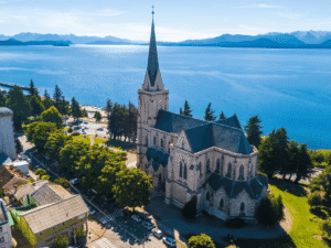 Historic church with tall steeple overlooking a lake and mountains in the background.