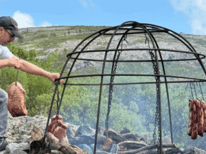 Grill dome with hanging meat over an open fire in outdoor setting.