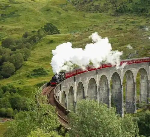 Vapor de tren antiguo cruzando un puente en un paisaje verde y montañoso.