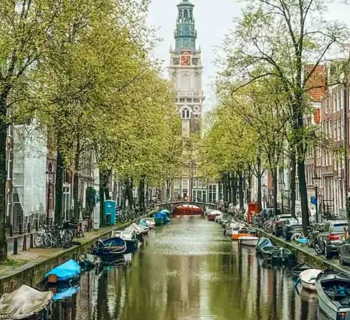 Puente sobre canal en Ámsterdam con edificios históricos y árboles verdes en primavera.